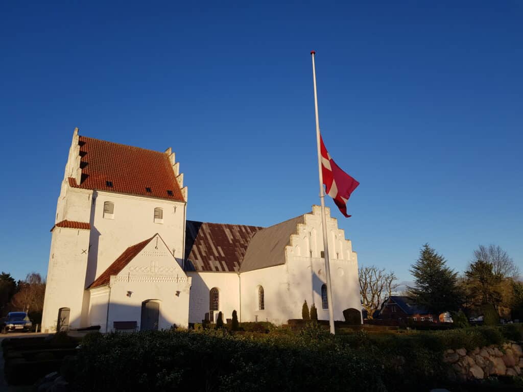 Sanderum Kirke med flag på halv og blå himmel ved bisættelse